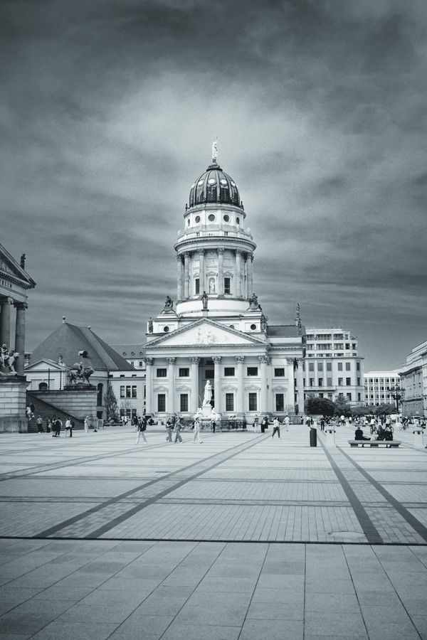 El empedrado de Gendarmenmarkt guía la mirada hasta la cúpula, con la gente pequeña bajo un cielo pesado.