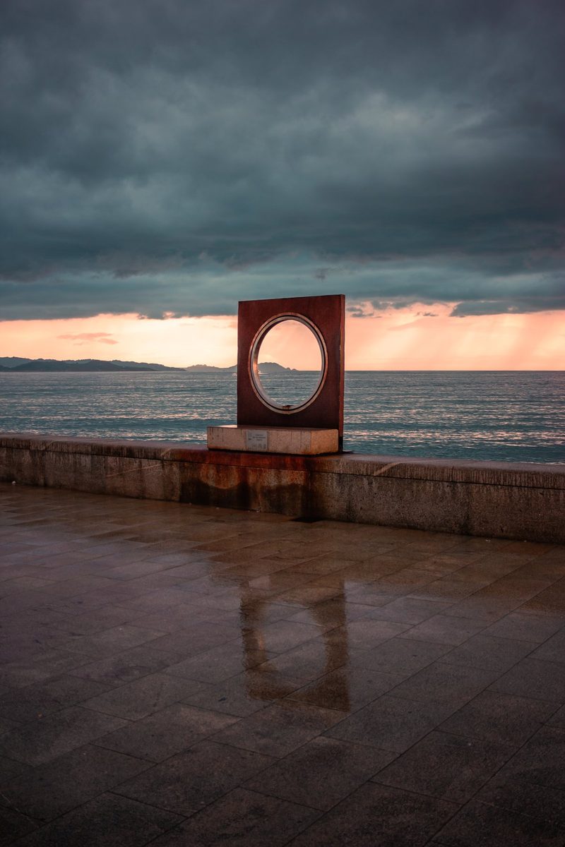 Un marco circular de acero sobre el paseo mojado, con el mar y un cielo de tormenta detrás.