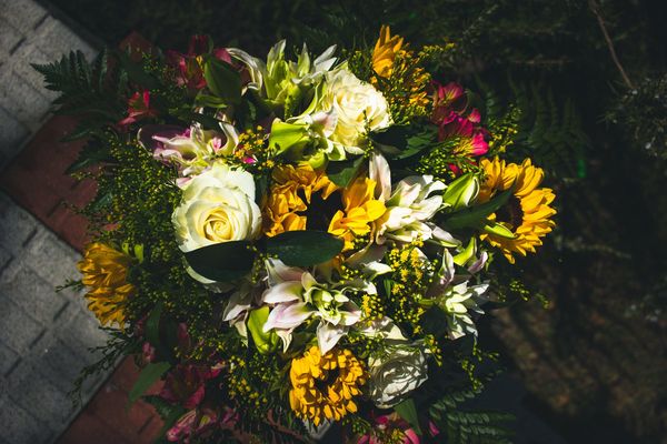 El mismo ramo visto desde arriba: rosas blancas, girasoles y alstroemerias rosas entre el verde, con la luz dura del mediodía.