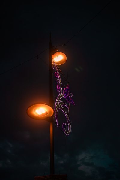 Una farola encendida con dos globos naranjas y una mariposa de luces violetas en la noche.