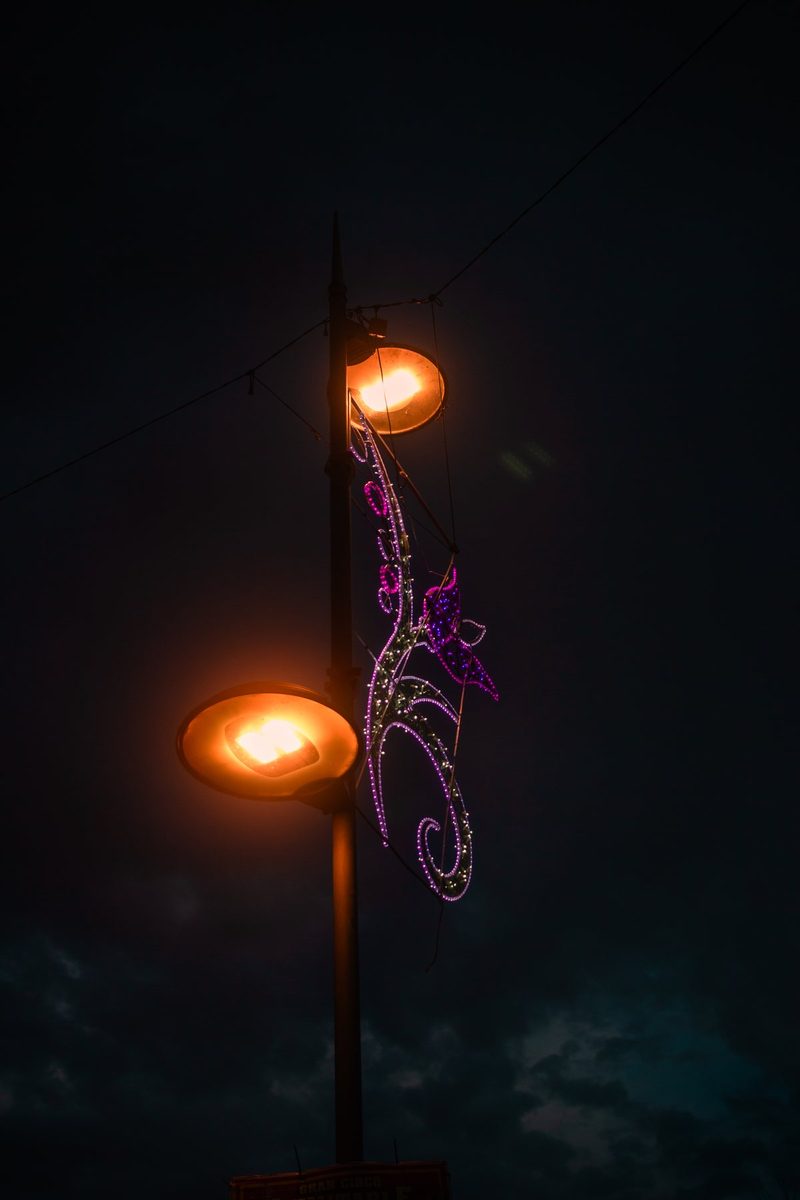 Una farola encendida con dos globos naranjas y una mariposa de luces violetas en la noche.