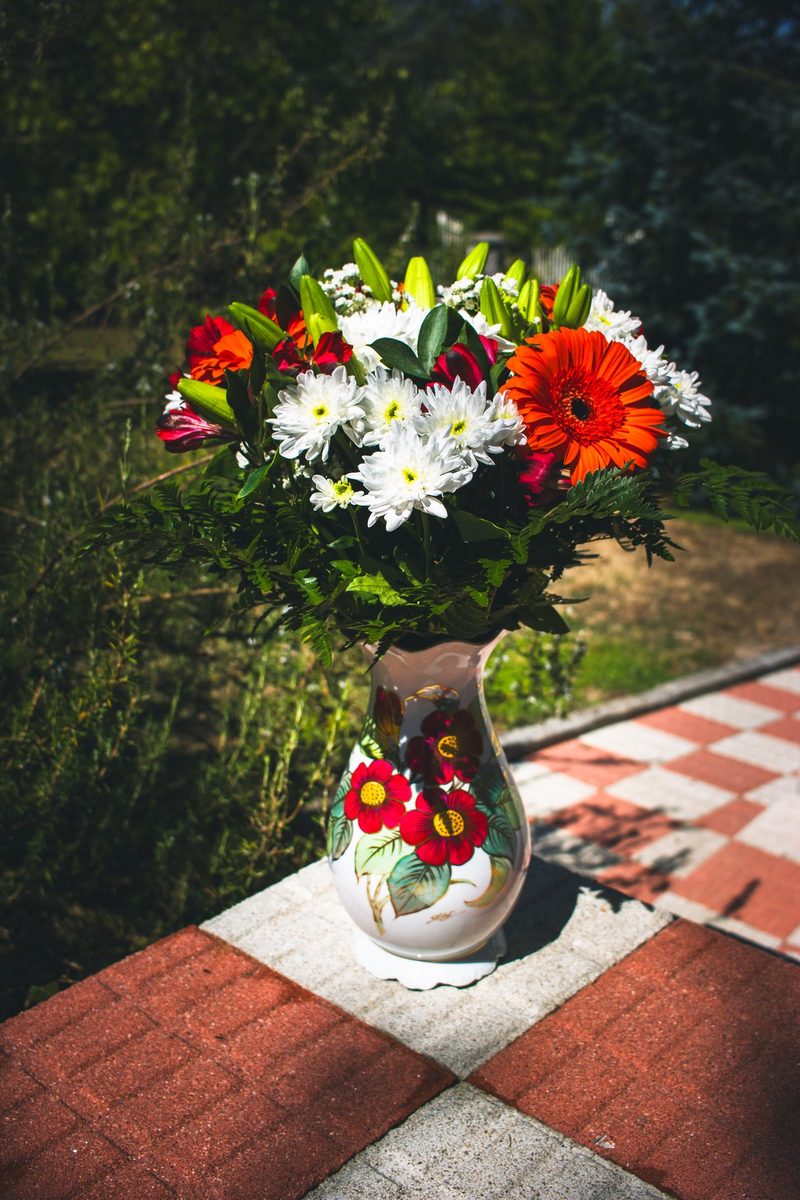 Un ramo de gerberas, crisantemos blancos y alstroemerias rojas en un jarrón pintado, sobre las baldosas al sol.