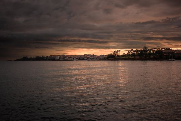 La ensenada en calma con Portonovo en el horizonte, entre un cielo oscuro y una línea de luz naranja.