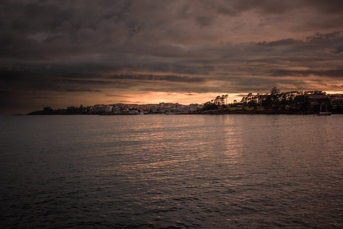 La ensenada en calma con Portonovo en el horizonte, entre un cielo oscuro y una línea de luz naranja.