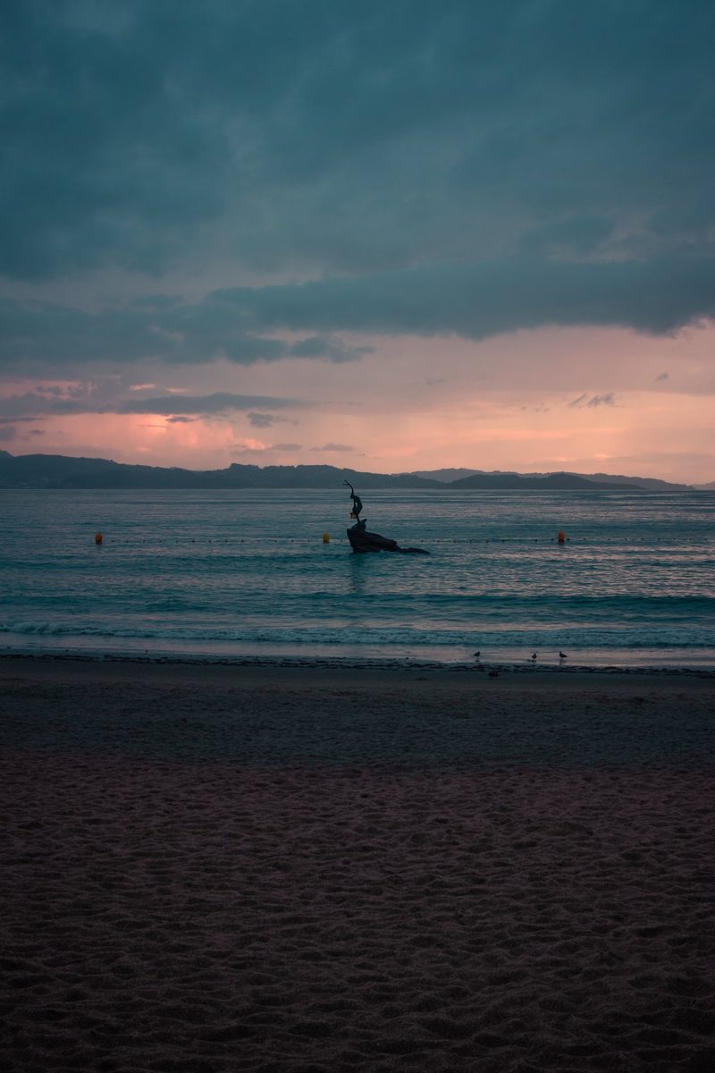 La Madama de Silgar sobre su roca en un atardecer azul, con una franja rosa sobre el horizonte.
