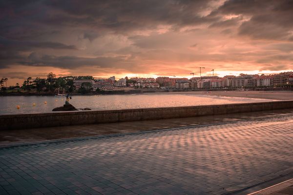 El paseo mojado en primer plano, la Madama de Silgar en el mar y la fachada de Sanxenxo al atardecer.