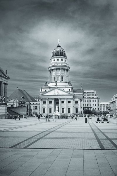 El empedrado de Gendarmenmarkt guía la mirada hasta la cúpula, con la gente pequeña bajo un cielo pesado.