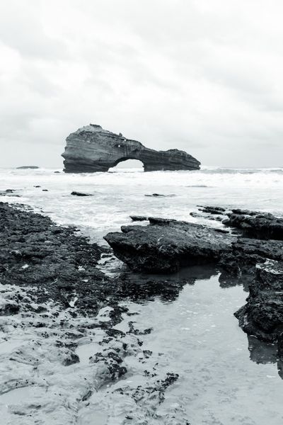 Una poza entre las rocas negras refleja el cielo; al fondo, el arco de piedra.