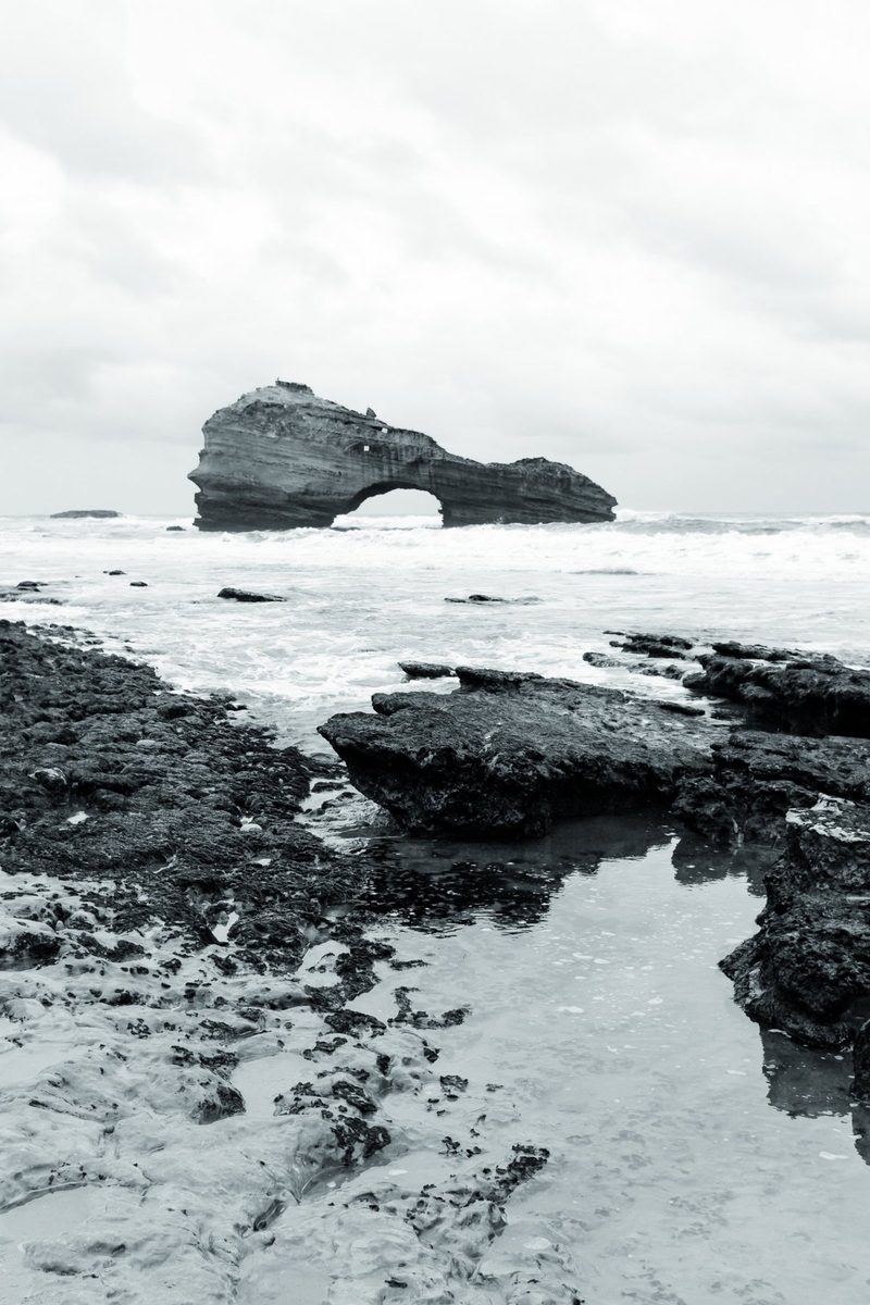 A pool among the black rocks reflects the sky; in the distance, the stone arch.
