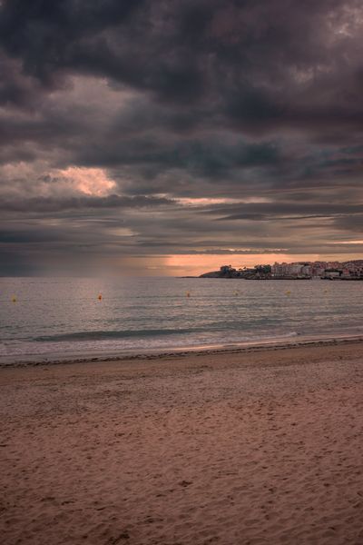 La arena mojada de Silgar, el mar en calma y Portonovo al fondo bajo un cielo de tormenta con una franja de luz.