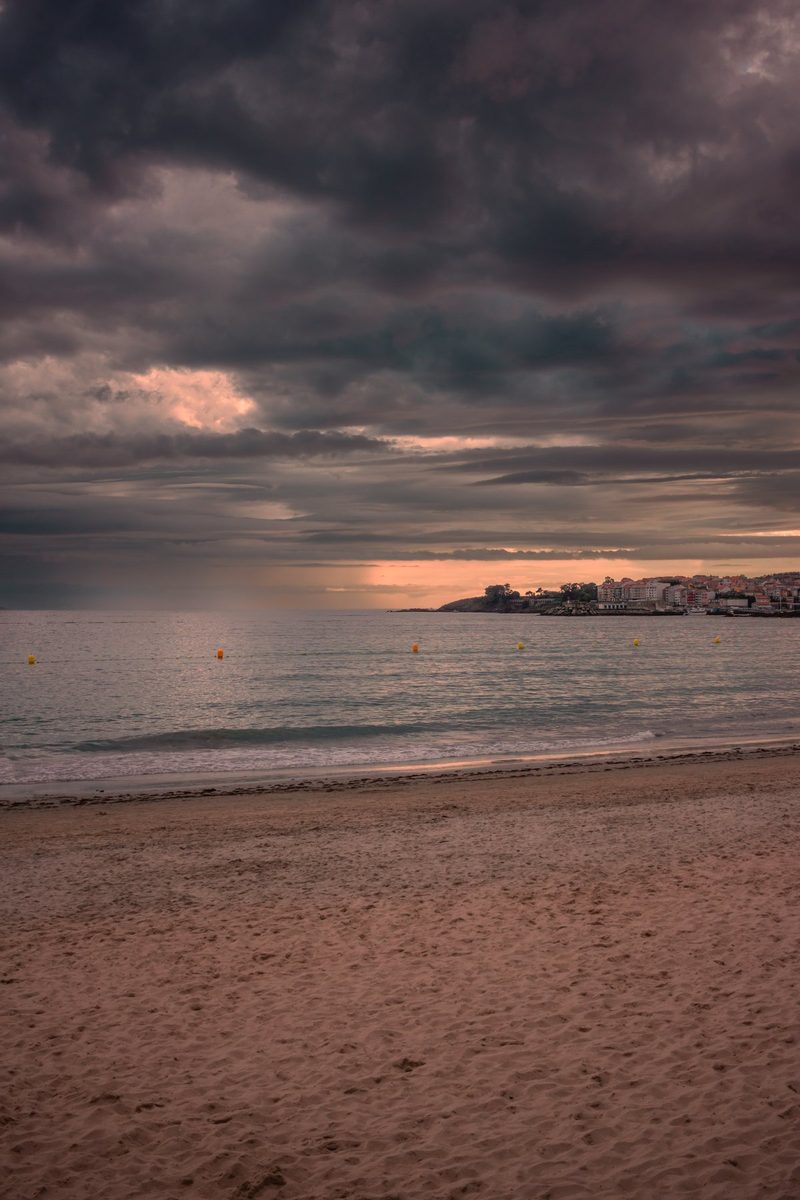 La arena mojada de Silgar, el mar en calma y Portonovo al fondo bajo un cielo de tormenta con una franja de luz.