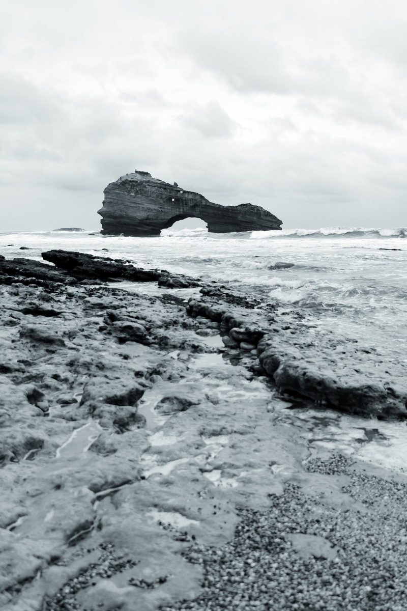 The rocks exposed at low tide lead the eye to the arch, under an overcast sky.