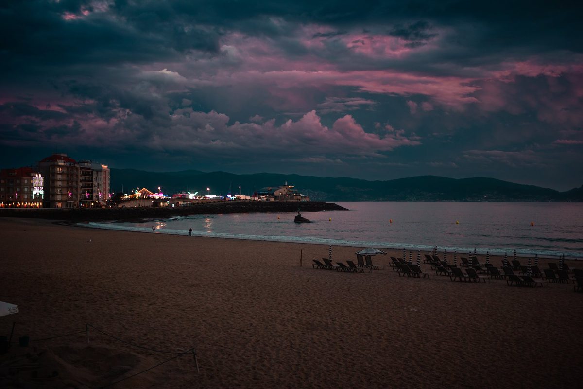 La playa de Silgar al anochecer, con las luces de la fiesta en el paseo y nubes rosas sobre la ría.