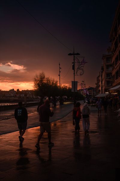 Siluetas de paseantes sobre el suelo mojado del paseo de Silgar, con el cielo naranja al fondo.