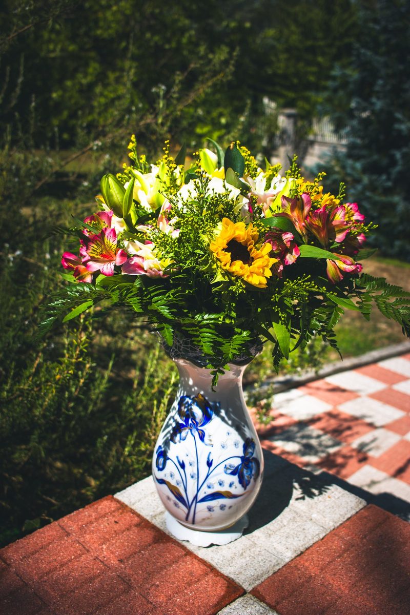Un ramo de girasoles, rosas y alstroemerias en un jarrón de cerámica azul y blanca, sobre las baldosas al sol.