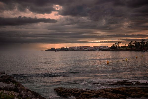 Rocas oscuras en primer plano, el agua quieta y Portonovo iluminado por el último sol bajo las nubes.