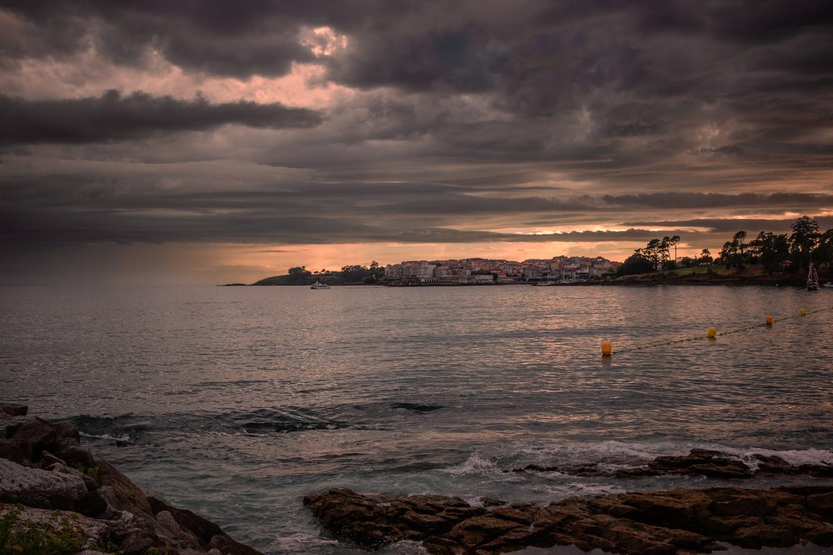 Rocas oscuras en primer plano, el agua quieta y Portonovo iluminado por el último sol bajo las nubes.