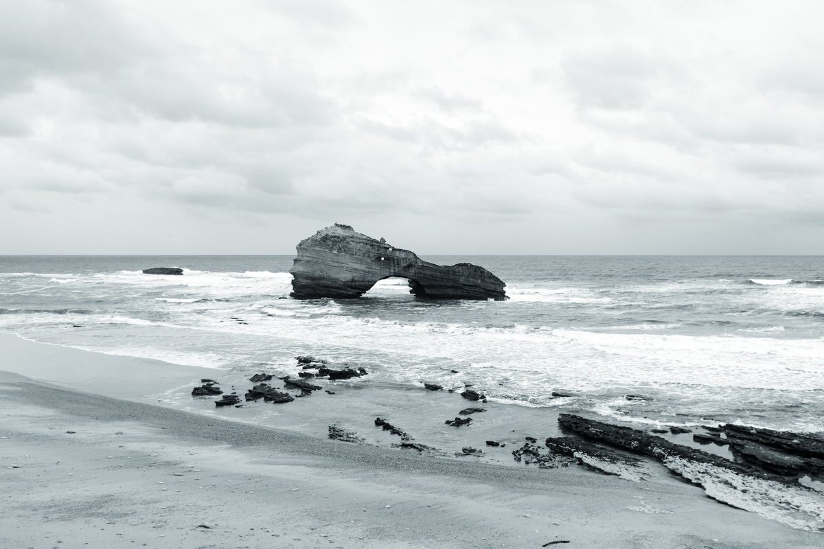 The Roche Percée in the middle of a rough sea, with the beach and rocks in the foreground.