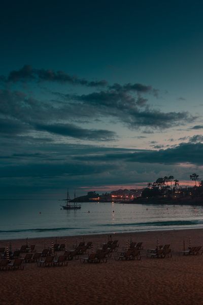 Un velero fondeado en la ensenada al anochecer, con las luces de la punta y las nubes azules sobre la ría.